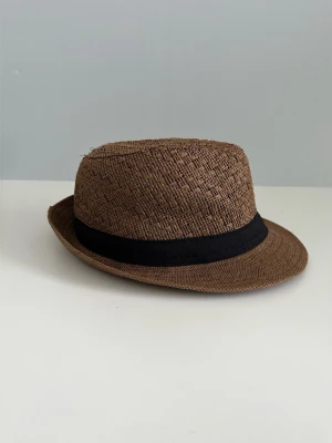 Brun stråhatt med svart band - Snygg brun stråhatt i storlek 60 med klassiskt svart band runt kullen. Hatten har ett flätat mönster och en kort brätte som ger en cool och avslappnad look. Perfekt accessoar för soliga dagar och festivaler.