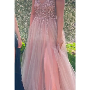 Rosa balklänning med paljetter - Underbar rosa balklänning med glittrande paljetter på överdelen och ett luftigt, flödande kjolparti. Klänningen har en v-ringad urringning och är ärmlös, vilket ger en elegant och festlig look. Perfekt för speciella tillfällen. Den är ganska stor till på överdelen, men går att sy in. 