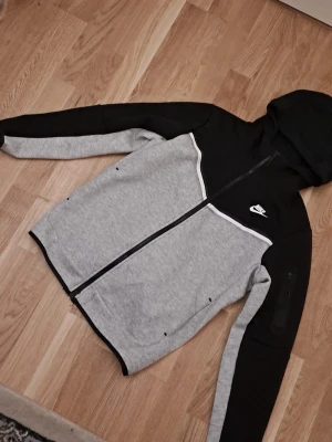 Nike Tech Fleece hoodie svart/grå - Nike Tech Fleece hoodie i svart och grått med dragkedja och huva. Snygg vit detalj över bröstet och klassisk Nike-logga på vänster sida. Mjukt och bekvämt material, perfekt för en sportig och avslappnad stil.