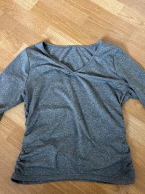 Grå v-ringad topp med rynkade sidor - Mjuk och stretchig grå topp med v-ringning och långa ärmar. Snygga rynkade detaljer längs sidorna ger en cool och trendig look. Perfekt att styla med jeans eller kjol för en avslappnad vibe.