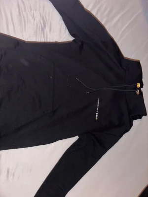 Svart hoodie Porsche x BOSS - Snygg svart hoodie från Porsche x BOSS med minimalistisk logga på bröstet. Hoodien har huva med dragsko och guldfärgade detaljer, samt en stor magficka framtill. Tillverkad i mjukt bomullsmaterial och har långa ärmar. Köpt från zalando.