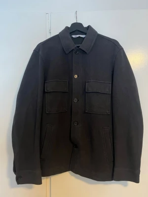 Mörkgrå overshirt från Arket - Stilren mörkgrå overshirt från Zara med klassisk krage och fyra fickor framtill, två med lock och två snedställda. Overshirten har raka ärmar med knapp vid ärmslut och stängs med knappar framtill. Perfekt lager på lager-plagg i ett kraftigare tyg.