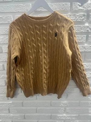 Beige kabelstickad tröja Polo Ralph Lauren - Kabelstickad tröja i beige från Polo Ralph Lauren med klassisk rund halsringning och broderad marinblå logga på bröstet. Tröjan har ribbade muddar vid ärmslut och nederkant. Perfekt för lager-på-lager och snygg till jeans.