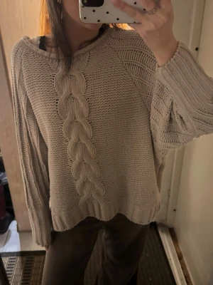 Beige stickad tröja med flätmönster - Mysig beige stickad tröja med breda flätor framtill och ribbade muddar. Tröjan har lång ärm och en relaxed, loose passform som ger en chill vibe. Perfekt att slänga på sig över ett linne eller t-shirt. Opps har ett litet hål syns på andra bilden 