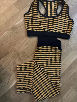 Orange och svart träningsset - Snyggt träningsset med sport-bh och tights i gult och svart mönster. Setet har breda svarta kanter och ett coolt grafiskt print. Perfekt för gymmet eller yoga. Materialet känns stretchigt och bekvämt.