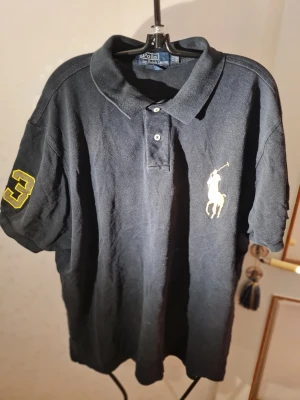Svart pikétröja från Ralph Lauren - Klassisk svart pikétröja från Ralph Lauren med stor gul logga på bröstet och siffran 3 på ärmen. Tröjan har krage, knappar framtill och är kortärmad. Perfekt för en stilren och sportig look.