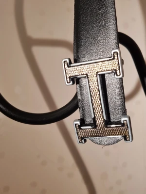 Svart bälte med metallspänne - Snyggt svart bälte i slätt läder med ett stort, silverfärgat metallspänne format som bokstaven I. Perfekt accessoar för att lyfta din outfit med en stilren detalj. Passar till både jeans och byxor.
