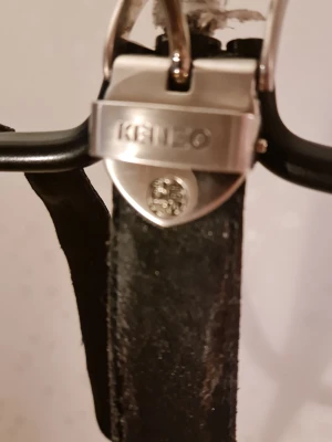 Svart Kenzo bälte i skinn - Snyggt svart bälte från Kenzo i äkta skinn med silverfärgat spänne där Kenzo-loggan är ingraverad. Bältet har en klassisk, smal design som passar till många olika stilar. Perfekt accessoar för att lyfta din outfit.