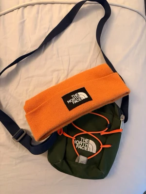  axelväska och mössa -  The North Face - Snygg grön axelväska från The North Face med justerbar marinblå rem och orange detaljer. Väskan har dragkedja både fram och bak samt en vit logotyp framtill. Tillverkad i slitstark textil, perfekt för att bära det viktigaste. North face mössa i samma klassiska orangea färg medföljer axelremsväskan. One size