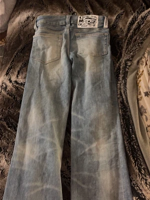 Ljusblå bootcut jeans från Cheap Monday - Säljer ett par ljusblå jeans från Cheap Monday med bootcut passform och klassiska bakfickor. Jeansen har en cool tvättad look och ett stort vitt märke bak i midjan. Perfekta för dig som gillar retrovibbar och avslappnad stil.