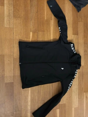 Svart vindjacka från Peak Performance - Sportig svart vindjacka från Peak Performance med dragkedja framtill och två sidofickor med dragkedja. Jackan har lång ärm med vit logotyptext längs ärmen och en liten logga på bröstet. Perfekt för aktiva dagar utomhus.