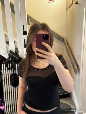 Svart topp med mesh och nitar H&M - Svart kortärmad topp från H&M med meshparti upptill och små silvriga nitar längs urringningen. Figurnära passform och stilren design som ger en edgy vibe.💖