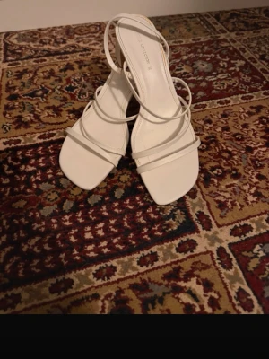 Vita strappy pumps med blockklack - Säljer ett par vita pumps från Pull&Bear med tunna remmar och fyrkantig tå. Skorna har en stabil blockklack och öppen häl, vilket ger en trendig och stilren look. Perfekta för dig som gillar minimalistisk design och vill ha något som sticker ut.