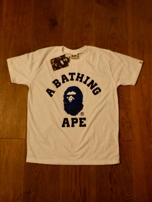 Vit A Bathing Ape t-shirt med tryck - Säljer en vit t-shirt från A Bathing Ape med klassiskt blått och svart logotryck på bröstet. T-shirten har rund hals, korta ärmar och är gjord i mjuk bomull. Perfekt för dig som gillar streetwear och vill sticka ut med en ikonisk design.