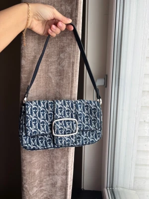 Mango blå axelremsväska med monogram - Snygg blå axelremsväska från MCM med vitt monogramtryck över hela väskan. Väskan har en rektangulär form, silverfärgat spänne framtill och justerbart axelband. Materialet är tyg med läderdetaljer. Perfekt för dig som gillar statement-accessoarer.