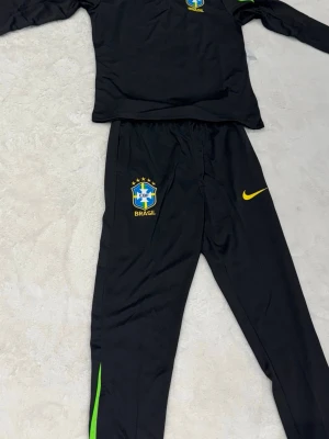 Svart Brasilien träningsset Nike - Snyggt svart träningsset från Nike med Brasilien-emblem på både tröja och byxor. Setet har gula detaljer och gröna inslag på byxbenen. Perfekt för fotbollsträning eller chill. Materialet är troligtvis polyester för bästa komfort och rörelsefrihet.