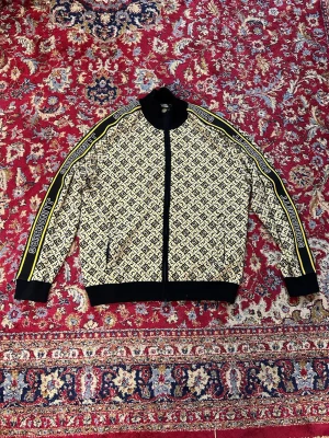 Beige och svart ziptröja J.Lindeberg - Säljer en beige och svart ziptröja från J.Lindeberg med grafiskt monogrammönster över hela plagget. Svarta muddar, krage och midja samt gula och svarta ränder med logga längs ärmarna. Tröjan har fickor med dragkedja och sportig look. Den mest komplimenterade och frågeskapande tröja jag haft. J.Lindeberg slog verkligen en home run med denna