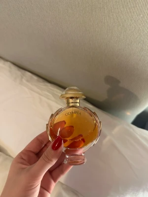 Olympea parfym från Paco Rabanne - Säljer en Olympea parfym från Paco Rabanne. Flaskan är rund med eleganta detaljer i guld och genomskinligt glas. Parfymen har en varm bärnstensfärgad vätska och flaskan har ett exklusivt och lyxigt utseende. Perfekt för dig som gillar unika dofter.