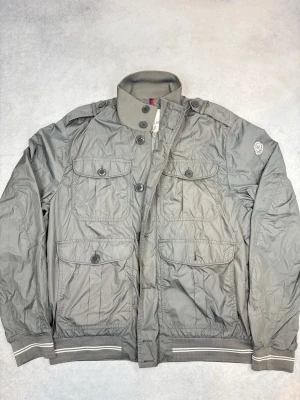 Grå vindjacka från Moncler - Stilren grå vindjacka från Moncler med fyra stora fickor framtill, hög krage och ribbade muddar. Jackan har knapp- och dragkedjestängning samt diskret logga på ärmen. Tillverkad i lätt och tåligt material, perfekt för blåsiga dagar.