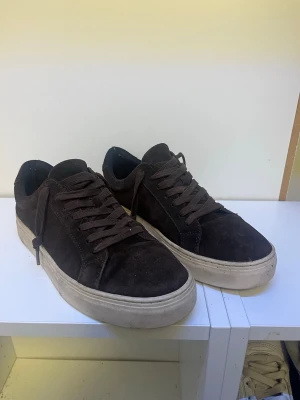 Mörkbruna vagabonds  - Snygga mörkbruna sneakers med ovandel i mocka och matchande bruna skosnören. Skorna har en vit, tjock sula som ger en cool kontrast till det mörka materialet. Perfekta för dig som gillar en stilren och enkel look.