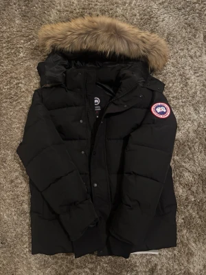 Canada Goose Vinterjacka - Säljer en svart dunjacka från Canada Goose med stor huva och avtagbar pälskant. Jackan har det klassiska Canada Goose-märket på ärmen och är fylld med dun för extra värme. Perfekt för kalla vinterdagar och har en stilren, tidlös design.