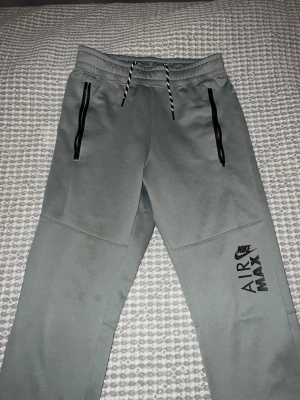 Grå Nike Air Max mjukisbyxor XS - Snygga grå mjukisbyxor från Nike Air Max med svarta dragkedjor på fickorna och justerbar dragsko i midjan. Tillverkade i mjukt Therma-FIT material som håller värmen. Perfekta för chill eller träning, med stor logga på benet.
