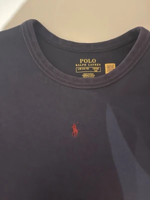 Svart t-shirt från Polo Ralph Lauren - Klassisk mörkblå sweatshirt från Polo Ralph Lauren med rund halsringning och den ikoniska röda broderade loggan på bröstet. Tillverkad i mjuk bomull som känns skön mot huden. Perfekt basplagg för en clean och stilren look.