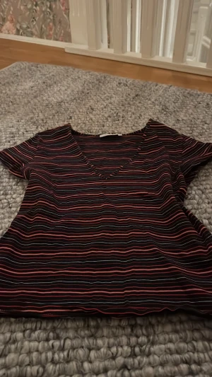 Randig V-ringad  - Kortärmad topp från Monki med V-ringning och smal passform. Svart bas med tunna ränder i rött, orange, rosa och blått. Tillverkad i mjukt och stretchigt material som sitter skönt på kroppen. Perfekt för en avslappnad och trendig look.