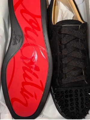 Svarta Louboutin sneakers med nitar - Säljer ett par svarta sneakers från Christian Louboutin med klassisk röd sula och svarta nitar på tån. Skorna har snörning, är tillverkade i mocka och har en lyxig vibe. Perfekta för dig som vill sticka ut med exklusiva detaljer och edgy look. 