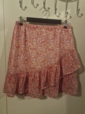 Blommig volangkjol i rosa toner - Supersöt kjol med volangkant och blommigt mönster i rosa, vitt och orange. Kjolen har en avslappnad passform med resår i midjan och är gjord i ett lätt, luftigt tyg. Perfekt för dig som gillar romantisk och feminin stil.