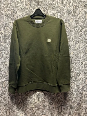 Olivgrön sweatshirt från Stone Island - Snygg olivgrön sweatshirt från Stone Island med rund halsringning och diskret logopatch på bröstet. Tröjan har långa ärmar, ribbade muddar och är tillverkad i mjuk bomull. Perfekt för en avslappnad och stilren look.