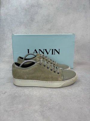 Grå sneakers från Lanvin - Snygga sneakers från Lanvin i grå mocka med matchande grå lackdetaljer på tån och klassisk vit sula. Skorna har grå skosnören och en stilren, låg profil som passar till många olika outfits. Perfekt för dig som gillar exklusiva och trendiga sneakers.