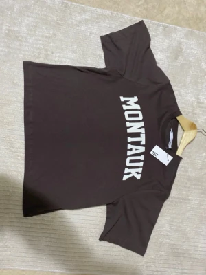 Brun Montauk t-shirt i bomull - Snygg brun t-shirt från Lager 157 med vit text 'MONTAUK' tryckt över bröstet. Perfekt till jeans eller shorts för en sommar outfit. Helt ny, inte använd alls. Ord.pris: 100kr. Meddela vid frågor! :) 