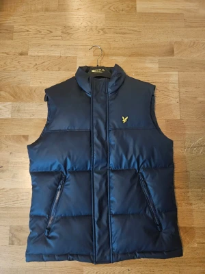 Mörkblå dunväst från Lyle & Scott - Snygg mörkblå dunväst från Lyle & Scott med gul logga på bröstet. Västen har hög krage, två dragkedjeförsedda fickor och quiltad design. Perfekt att slänga över en hoodie eller tröja för extra värme.