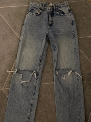 Ljusblå jeans - Säljer ett par ljusblå mom jeans med hög midja och slitningar över knäna. Jeansen har raka ben. Perfekta för en avslappnad och trendig look. Materialet är denim i bomull.