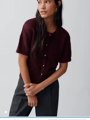 Vinröd kortärmad kofta med pärlknappar - Mysig vinröd kofta med korta ärmar och rund halsringning. Koftan har en fluffig, stickad struktur och stängs med små pärlknappar. Knappt använd 