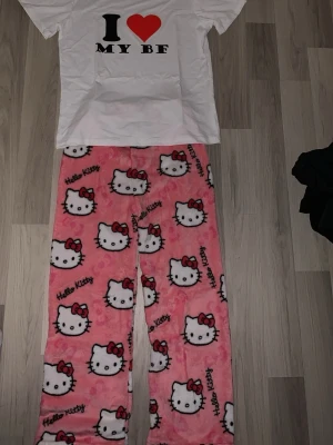 Hello Kitty pyjamas  - Supergullig pyjamas med vit t-shirt med texten 'I ❤️ MY BF' och rosa fluffiga byxor med Hello Kitty-motiv och röd rosett. Perfekt för mysiga hemmakvällar. Byxorna är i mjukt material och har ett lekfullt mönster.