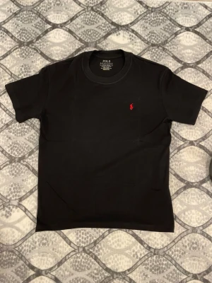 Svart t-shirt från Polo Ralph Lauren - Svart t-shirt från Polo Ralph Lauren med klassisk rund hals och korta ärmar. T-shirten har en liten röd broderad logga på bröstet och är tillverkad i mjuk bomull för en skön känsla. Perfekt för en stilren och enkel look.