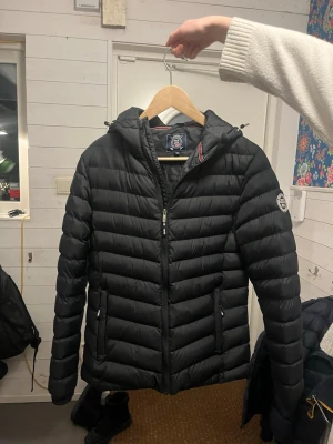 Svart pufferjacka med huva från Polo Club  - Snygg svart pufferjacka från Polo Club med huva och dragkedja framtill. Jackan har två sidofickor med dragkedja och ett märke på sidan av armen. Jackan är i storlek Junior M 