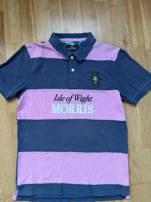 Randig pikétröja från Morris - Säljer en snygg pikétröja från Morris med breda rosa och mörkblå ränder. Det står att skjortan är L men den passar S Skjortan har krage, knappar framtill och broderad logga på bröstet. Materialet är mjuk bomull och insidan av kragen har ett blommigt tyg. Perfekt för en avslappnad men stilren look.