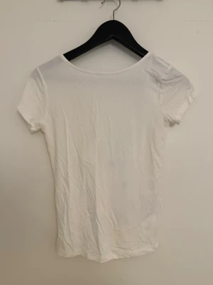 Vit kortärmad topp med knyt - Säljer en vit kortärmad topp med djup v-ringning och snygg knytning där bak. Toppen är i mjukt material och har en enkel, clean look som passar till mycket. Perfekt för dig som gillar minimalistisk stil med en twist.