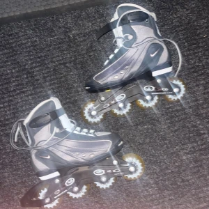 Svarta inlines från Nike - Säljer ett par svarta och grå inlines från Nike med vita detaljer. De har snörning, spänne upptill och gula genomskinliga hjul. Snygg sportig design och robust ram i metall och plast. Perfekta för dig som gillar att åka snabbt och smidigt.