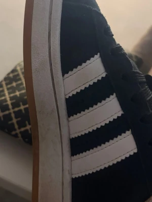 Svarta Adidas sneakers med vita ränder - Klassiska svarta Adidas sneakers med tre vita ränder på sidan och brun sula. Skon har en låg profil och ovandelen verkar vara i mocka. Perfekt för dig som gillar en tidlös och sportig stil.
