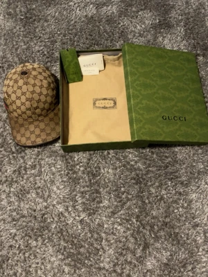 Gucci beige GG  - Säljer en beige keps från Gucci i klassiskt GG Supreme-mönster. Kepsen har en böjd skärm och ikoniska gröna och röda ränder på sidan. Tillverkad i canvasmaterial och levereras med originalkartong och dustbag. Säljer billigare pågrund av defekt i spännet