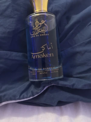 Oud El Emrat Amaken Eau de Parfum - Snygg parfymflaska från Oud El Emrat med namnet Amaken. Flaskan är genomskinlig med blått innehåll och har gulddetaljer på korken. Rymmer 100 ml och har en elegant, modern design med arabiska och engelska texter på framsidan.