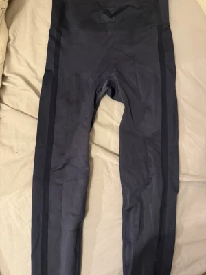 Svarta leggings från Puma x Vogue - Snygga svarta leggings från Puma x Vogue med diskreta mörka ränder längs benen. Hög midja och tight passform, perfekta för träning eller en sportig look. Tillverkade i ett stretchigt syntetmaterial som sitter skönt hela dagen.