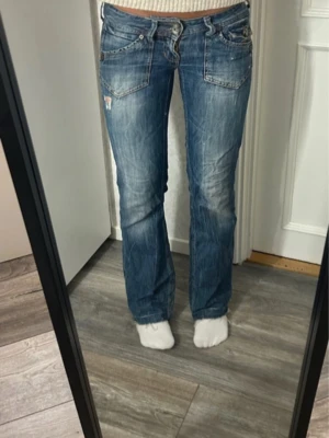 Blå bootcut jeans från EDC - Säljer ett par blå bootcut jeans från EDC med slitningar och snygga detaljer på bakfickorna. Jeansen har låg midja, fem fickor och klassisk knappgylf. Materialet är denim i bomull med en cool tvättad look och broderade detaljer på fickorna. Midjemått 40 innerben 80