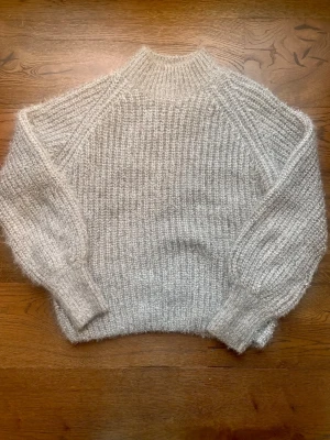 Silvrig fluffig stickad tröja - Mysig silvrig stickad tröja med hög hals och breda ribbstickade muddar. Tröjan har en fluffig struktur och är tillverkad i ett mjukt material, perfekt för kyliga dagar. 