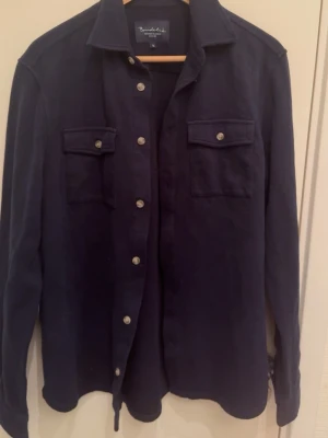 Mörkblå overshirt från M•Q - Stilren mörkblå overshirt från  M•Q med klassisk krage, tryckknappar och två bröstfickor med lock. Tillverkad i mjukt bomullsmaterial och har långa ärmar. Perfekt lager-på-lager-plagg för en avslappnad look.