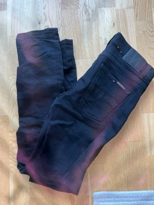 Svarta jeans från Nudie Jeans - Säljer ett par svarta jeans från Nudie Jeans i modellen 32/34. Klassisk femficksdesign med svarta sömmar och läderpatch bak i midjan. Snygg passform och perfekt basplagg till garderoben. Materialet är mjukt och stretchigt för extra komfort.